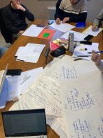 Une table de travail lors d'une réunion. Il y a des papiers et un ordinateur, ainsi que des gens autour de la table, en train de réfléchir.